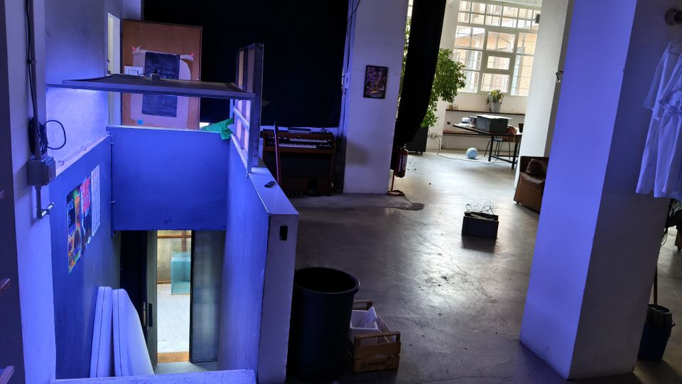 Interno di un loft o studio industriale con pavimento in cemento e pareti bianche, con un mezzanino e una scala che scende a un livello inferiore. Sullo sfondo grandi finestre a griglia illuminano una zona con tavolo e attrezzature, con colonne, tende nere e oggetti sparsi nello spazio.