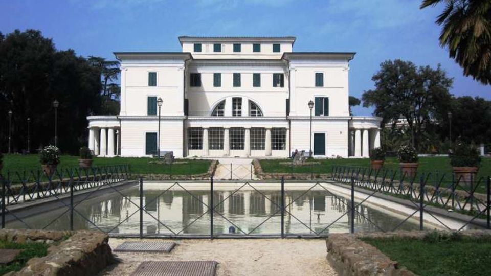 Facciata frontale di una villa storica bianca a più piani con finestre regolari e un grande arco centrale, affiancata da porticati con colonne. In primo piano si vede una vasca rettangolare con acqua e recinzione metallica, circondata da prato e alberi.