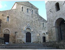 Facciata di una chiesa romanica in pietra con portale ad arco e piccole finestre strette, affacciata su una piazza con gradini in pietra. A destra si vede una struttura adiacente con grandi arcate e una cancellata in ferro.