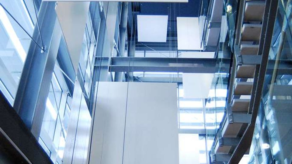 Atrio interno di un edificio moderno con pareti vetrate e struttura in acciaio, illuminato da luce naturale dall’alto. Grandi pannelli bianchi sospesi al soffitto scendono verticalmente nello spazio centrale sopra passerelle e scale in vetro.