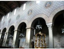 Interno di una chiesa con navata laterale scandita da grandi archi su colonne, soffitto ligneo e lampadari sospesi. Sulla parete in muratura sono presenti finestre alte e medaglioni decorativi, con un baldacchino dorato in primo piano.