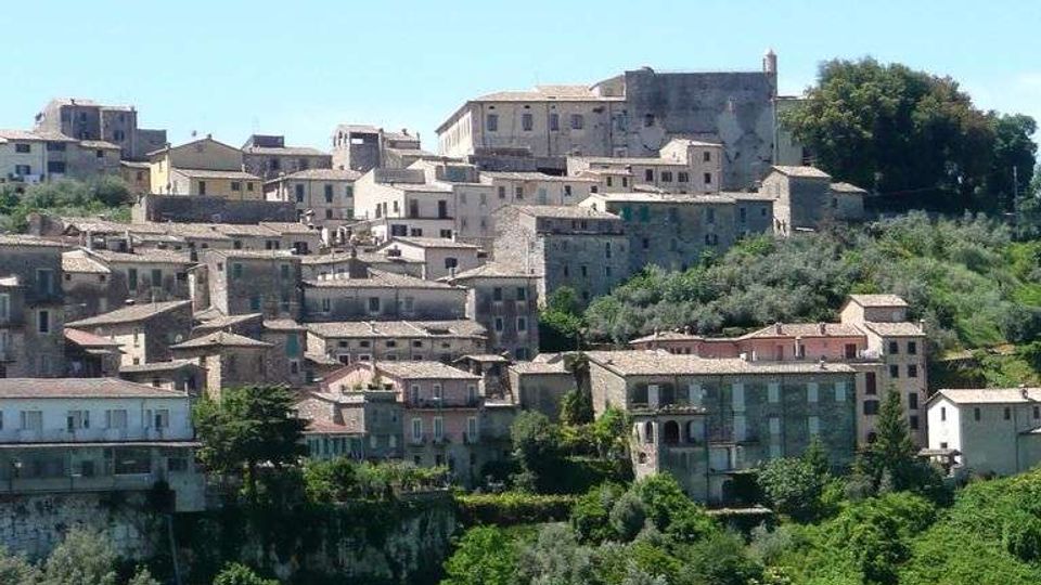 Panorama di un borgo collinare con case in pietra e tetti in coppi, addossate su diversi livelli lungo il pendio. In alto si distingue un grande edificio storico, mentre in primo piano si estende una vallata verde con alberi e vegetazione fitta.