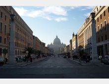 Lungo viale urbano rettilineo con carreggiata ampia, fiancheggiato da edifici storici e lampioni monumentali, che conduce in prospettiva verso la cupola della Basilica di San Pietro sullo sfondo. Sul bordo della strada si notano marciapiedi, alcune palme e auto parcheggiate.