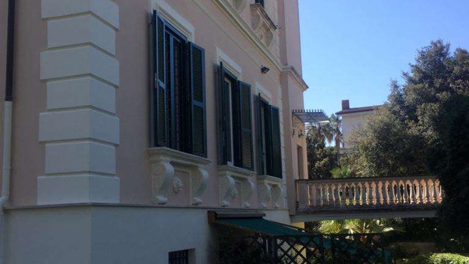 Facciata laterale di una villa storica intonacata color crema, con persiane scure e cornici in rilievo alle finestre. In primo piano si vede un cortile pavimentato in pietra con vaso ornamentale e, sullo sfondo, una balaustra e un giardino con alberi.