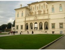 Facciata di un grande palazzo storico in stile classico, con decorazioni in rilievo, finestre alte e una loggia ad archi al piano terra. Davanti all’ingresso centrale con scalinata e balaustra si estende un ampio prato verde, con vialetti e alberi sullo sfondo.