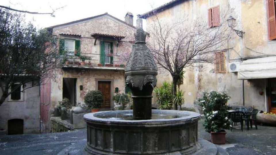 Piazzetta di un borgo con una fontana in pietra al centro su basamento circolare, circondata da edifici intonacati e invecchiati con persiane in legno e balconcini. Intorno si vedono vasi con piante, un albero spoglio e un piccolo spazio con tavolini all’aperto.