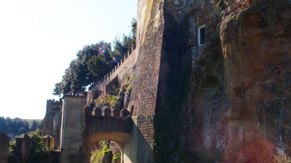 Ingresso ad arco in muratura lungo una strada in salita, ai piedi di una scogliera rocciosa con alti muri di contenimento. Sopra l’arco si vedono edifici storici in pietra con finestre e un camminamento fortificato che segue il bordo della rupe.
