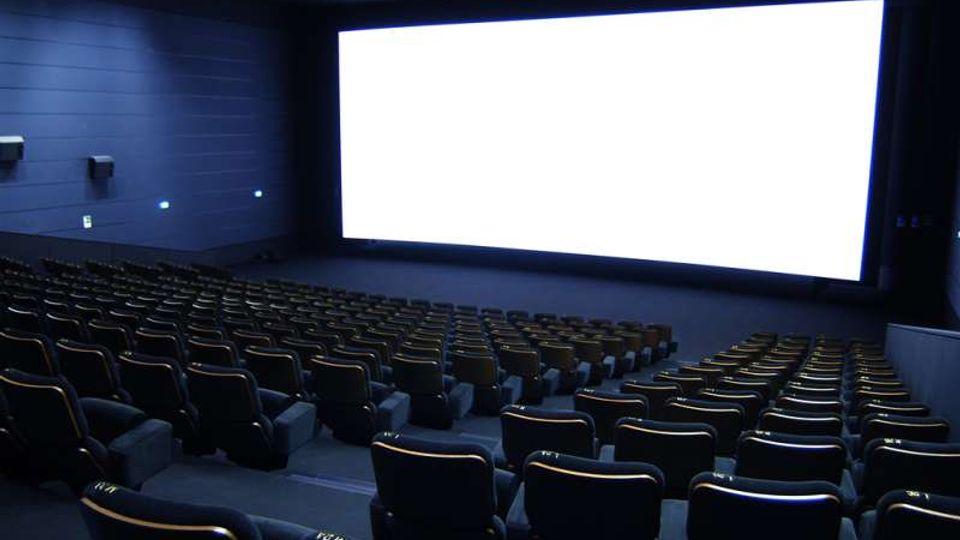 Sala cinematografica con file di poltrone scure disposte in gradinata e rivolte verso un grande schermo luminoso. Pareti e soffitto scuri con illuminazione discreta laterale e altoparlanti montati sulle pareti.