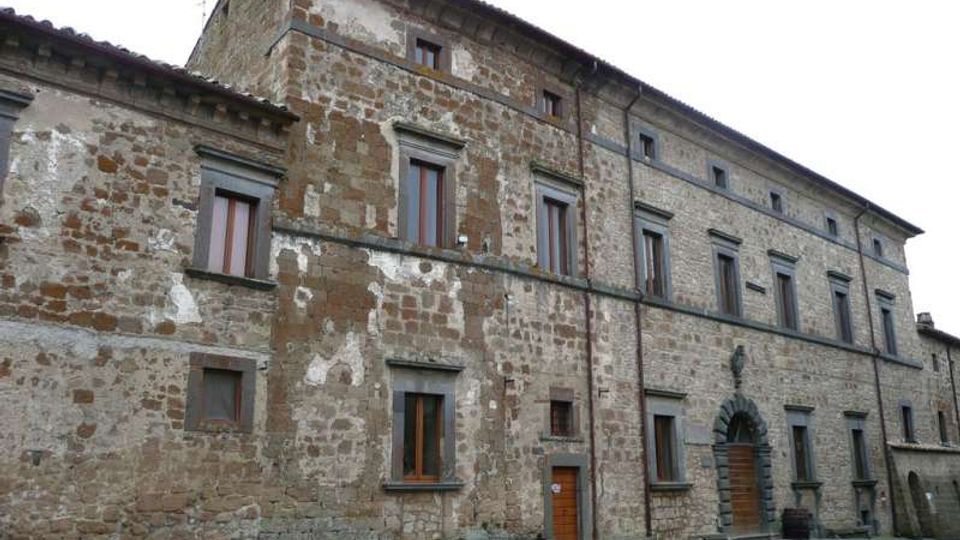 Edificio storico in pietra con facciata irregolare e intonaco consumato, disposto su più piani con finestre rettangolari incorniciate e tetto a falde. Sul lato destro si vede un grande portale ad arco in pietra che si affaccia su una strada lastricata.