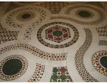Pavimento decorativo in mosaico con grandi motivi circolari e ottagonali intrecciati, realizzati con tessere geometriche multicolori. La superficie chiara è intervallata da fasce e rosoni con pattern ripetuti in verde, rosso, giallo e nero.