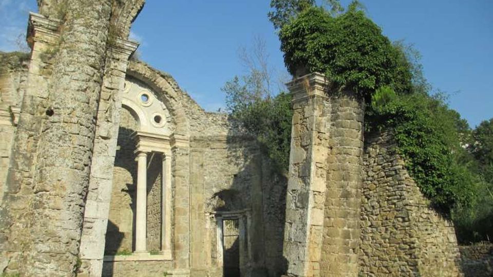 Rovine di un edificio storico in pietra, con facciata parzialmente crollata e un arco con colonne e piccole finestre circolari. Alti pilastri e muri perimetrali rimasti in piedi sono coperti in parte da vegetazione, in un’area erbosa sotto cielo sereno.
