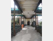 Passaggio pedonale coperto con pavimentazione a mosaico, colonne metalliche e ringhiere decorative, che conduce all’ingresso ad arco di un edificio in mattoni e intonaco. La copertura mostra segni di usura e ai lati si intravedono alberi e strutture schermate con teli.