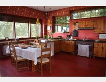 Cucina rustica con pavimento in cotto rosso, pareti con carta da parati rossa e mobili in legno con piano di lavoro ad angolo e fornelli. Al centro c’è un tavolo da pranzo con tovaglia bianca e sedie in legno, vicino a grandi finestre che affacciano su alberi all’esterno.