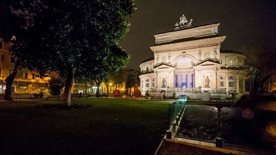 Veduta notturna di un edificio monumentale in stile neoclassico, illuminato, con grande facciata simmetrica e frontone decorato, affacciato su un giardino pubblico. In primo piano si vede un percorso con parapetto in vetro, alberi e prato che circondano la piazza.