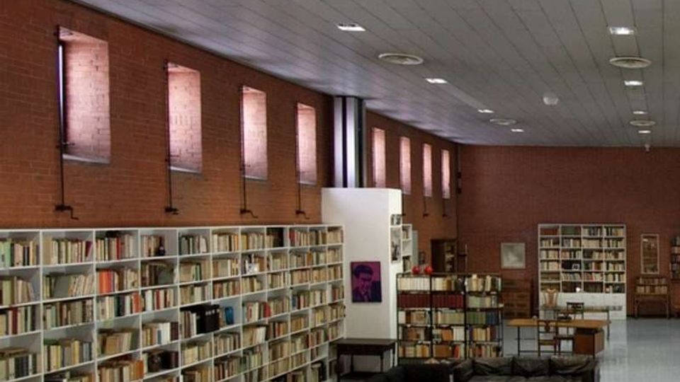 Interno di una biblioteca o sala lettura con pareti in mattoni rossi, alte finestre strette e soffitto a pannelli con illuminazione a incasso. Lungo la parete si estendono scaffali bianchi pieni di libri, con un’area lounge centrale con divani scuri, tavolini e tappeto.