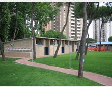 Edificio basso in muratura con tetto inclinato e porte blu, situato in un parco con prato verde, alberi alti e un sentiero pavimentato curvo in primo piano. Sullo sfondo si vedono alti palazzi residenziali e alcuni arredi esterni coperti.