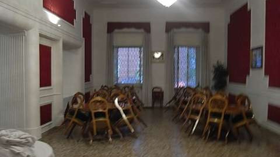 Sala interna ampia con pavimento in terrazzo, pareti bianche con pannelli rossi e un grande lampadario centrale. Ai lati sono impilate sedie in legno e sul fondo ci sono due finestre con tendine e una piccola applique a parete.