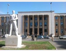 Edificio pubblico in stile razionalista con facciata simmetrica, alto portico a colonne e la scritta “IVSTITIA” sopra l’ingresso, affacciato su una piazza con auto parcheggiate. In primo piano si vede una grande statua bianca su piedistallo in un’area verde.