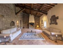Interno di un soggiorno rustico con pareti in pietra a vista, soffitto con travi in legno e pavimento in cotto. Al centro c’è un camino acceso con ampia cappa, affiancato da due divani chiari, un tappeto decorato e un tavolino in ferro, con nicchie e finestre incassate nelle mura.
