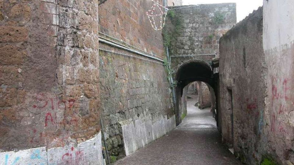 Vicolo stretto in pietra con pavimentazione in ciottoli, fiancheggiato da alti muri in muratura grezza con tubazioni a vista e tracce di graffiti. In fondo si vede un arco in pietra che forma un passaggio coperto verso una piccola curva della strada.