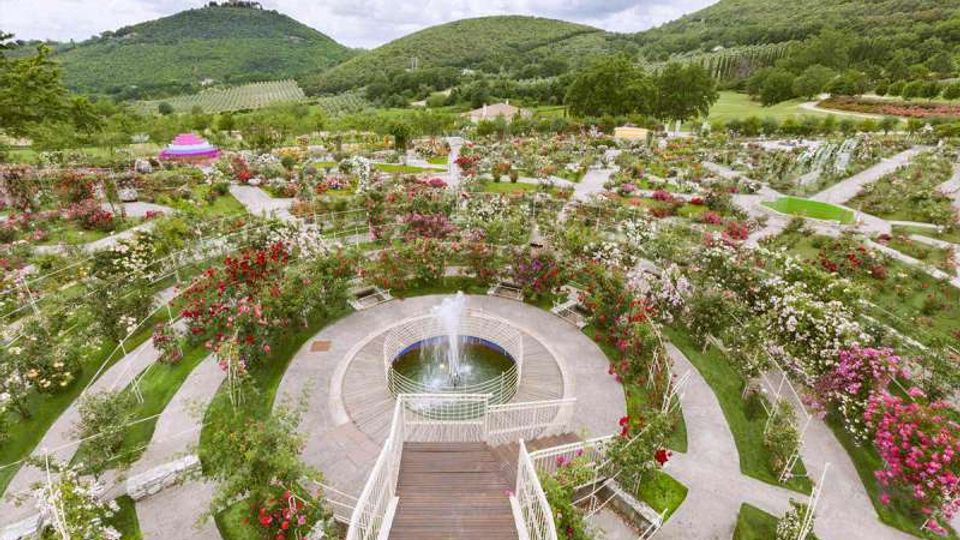 Giardino paesaggistico con aiuole fiorite e pergolati, organizzato in vialetti curvi attorno a una fontana circolare centrale con getto d’acqua. Sullo sfondo si vedono colline verdi e campi coltivati, con alberi e piccoli edifici sparsi nel paesaggio.