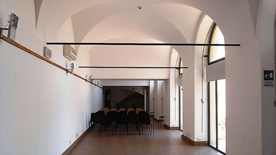 Interno di una sala rettangolare con soffitto a volte bianche e pavimento in mattoni, illuminata da grandi porte-finestre ad arco sul lato destro. In fondo si vedono file di sedie disposte come per una riunione o conferenza.