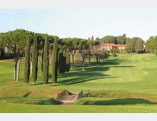 Ampio campo da golf con prati ondulati e un piccolo ponte in legno in primo piano. Lungo il percorso si vedono filari di cipressi e pini a ombrello, con edifici rurali dal tetto rosso sullo sfondo.