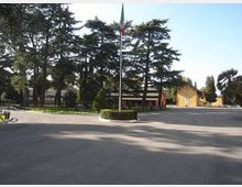 Ampio piazzale asfaltato con aiuola circolare e alto pennone centrale con bandiera. Sullo sfondo si vedono edifici bassi in stile caserma o complesso scolastico, circondati da alti alberi sempreverdi e aree verdi.