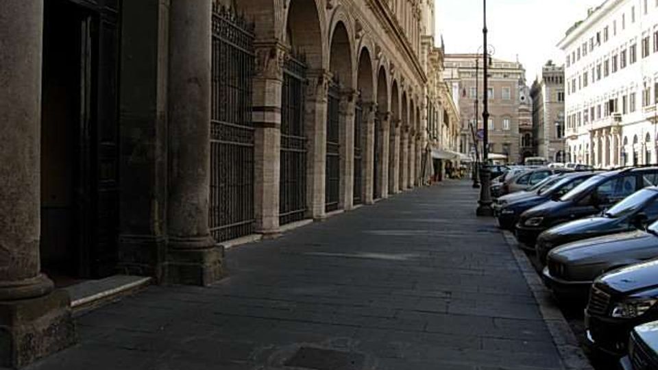 Portico urbano con lunga fila di arcate in pietra e colonne su un ampio marciapiede lastricato, affiancato da una strada con auto parcheggiate e lampioni. Sullo sfondo si vedono edifici storici e una piazza all’incrocio.