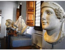 Interno di una sala museale con una fila di busti scultorei in pietra esposti su piedistalli lungo un corridoio. Sullo sfondo si vedono finestre con grata e persiane in legno, pareti chiare e pavimento in legno.