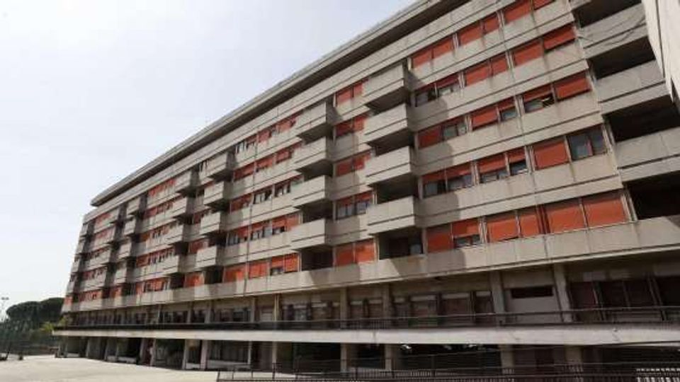 Grande edificio residenziale a più piani in cemento, con lunghe file di finestre e balconi e pannelli frangisole rossi. La struttura è sollevata su pilastri con un livello inferiore porticato e una passerella lungo la facciata.