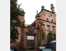 Edificio storico in mattoni con facciata alta e finestre incorniciate, con elementi architettonici scuri e un piccolo torrione sul tetto. In primo piano si vede un ingresso monumentale con pilastri e cancello, un’insegna, vegetazione rampicante e auto parcheggiate lungo la strada.
