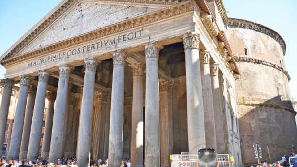 Facciata di un tempio romano con alto portico a colonne corinzie e frontone triangolare in pietra, con iscrizione latina. Sullo sfondo si vede la grande rotonda in muratura della struttura, in una piazza urbana.