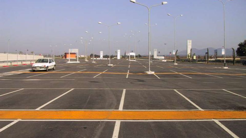 Ampio parcheggio asfaltato quasi vuoto, con stalli segnati da linee bianche e file di alti lampioni. Sullo sfondo si vedono recinzioni, alcuni cartelli e colline lontane sotto un cielo sereno.