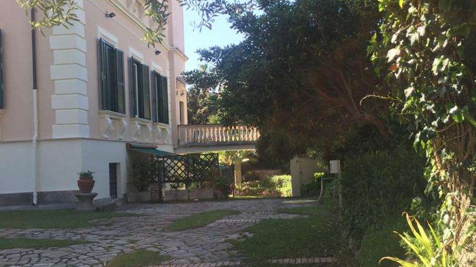 Vialetto in pietra e mattoni che attraversa un giardino alberato accanto a una villa in stile classico con facciata rosa e persiane verdi. Vegetazione fitta e alberi ombreggiano il percorso, con una balaustra e terrazza visibili sullo sfondo.