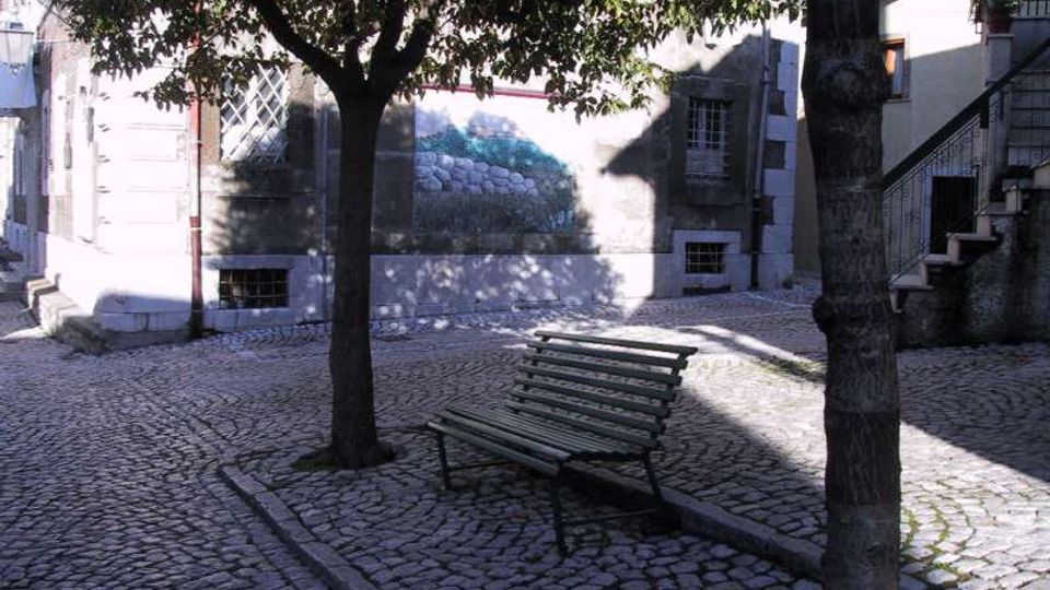 Piccola piazzetta con pavimentazione in ciottoli, una panchina metallica e alberi che creano ombra. Sullo sfondo si vedono facciate di edifici in pietra con finestre grigliate, una scala esterna e un murale dipinto sul muro.