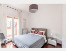 Camera da letto luminosa con letto matrimoniale, comodini con lampade e pavimento in legno; grandi porte finestre con tende bianche si aprono su un piccolo balcone. Pareti bianche con nicchie a muro, radiatore e lampadario a soffitto.