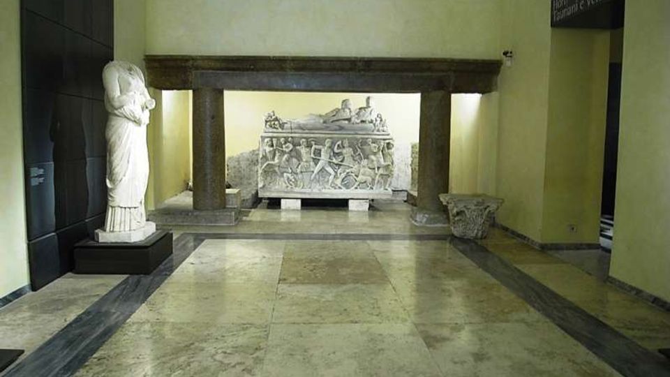 Sala museale con pavimento in marmo e pareti chiare, con al centro una struttura in pietra a colonne che incornicia un grande sarcofago scolpito in rilievo. A sinistra è esposta una statua su piedistallo e a destra si vede l’ingresso verso un’altra area espositiva.