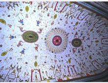 Soffitto decorato con affreschi su fondo chiaro, ricco di motivi geometrici e floreali e di figure stilizzate disposte in modo radiale attorno a grandi medaglioni circolari colorati. Lungo i bordi corre una fascia ornamentale con disegni ripetuti e simboli.
