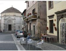 Piccola piazza con pavimentazione in pietra e tavolini metallici all’aperto davanti a edifici storici con balconi e fioriere. Sullo sfondo si vede una chiesa in pietra chiara con facciata decorata e tetto ottagonale, con auto parcheggiate lungo la strada.