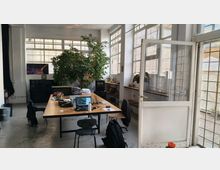 Interno di un ufficio open space in stile industriale con grandi finestre a griglia e porta vetrata, illuminato da luce naturale. Al centro c’è un lungo tavolo da lavoro con laptop e sedie, affiancato da una grande pianta in vaso e da mobili e scaffali lungo le pareti.