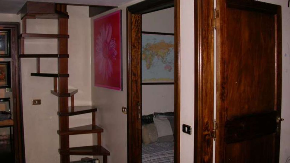 Interno di un’abitazione con scala a giorno in legno scuro e gradini sospesi lungo la parete. Accanto si vedono porte e telai in legno, una porta aperta, e una parete con quadri decorativi e una mappa.