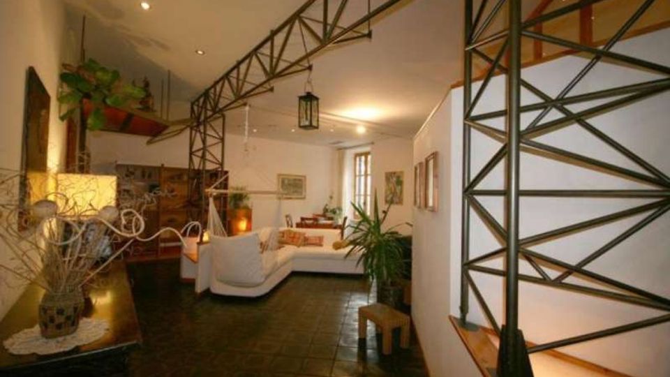 Interno di un soggiorno open space con pavimento scuro piastrellato, divani chiari e arredi in legno, illuminato da faretti a soffitto e una finestra sul fondo. Una struttura in metallo a traliccio separa parzialmente gli ambienti e si affaccia su una scala, con piante e quadri alle pareti.