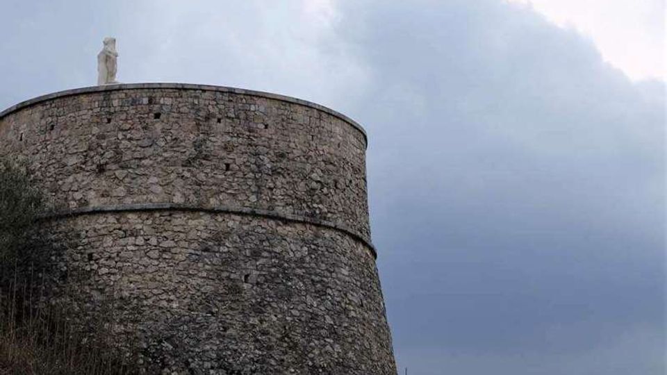 Torre fortificata cilindrica in pietra, con fascia orizzontale a metà altezza e una piccola statua sulla sommità. La struttura si staglia contro un cielo nuvoloso, con vegetazione bassa alla base e colline sullo sfondo.
