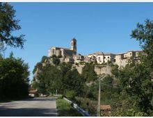 Borgo storico su uno sperone roccioso, con case in pietra e una chiesa con campanile al centro, circondato da alberi e mura di contenimento. In primo piano una strada asfaltata con guardrail e cavi elettrici sotto un cielo sereno.