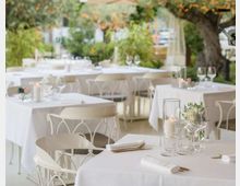 Terrazza di ristorante all’aperto con tavoli quadrati coperti da tovaglie bianche, apparecchiati con bicchieri e candele. Sedie bianche in metallo e vegetazione circostante con alberi e luci decorative sullo sfondo.