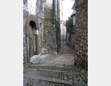 Vicolo stretto in un borgo storico con pavimentazione in ciottoli e mattoni, in lieve discesa tra edifici in pietra e intonaco segnati dal tempo. Si vedono una porta ad arco, un lampione a parete e muri ravvicinati che incanalano la strada.