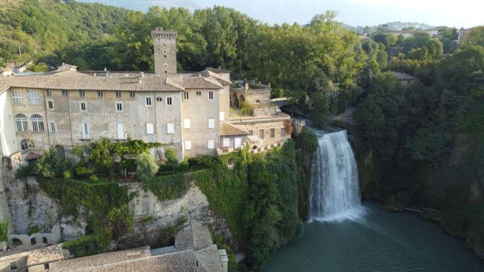 Veduta aerea di un edificio storico in pietra con torre quadrata e tetti in coppi, costruito su una rupe boscosa. Accanto all’edificio scende una cascata alta in un bacino d’acqua, circondato da alberi e colline.