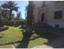 Giardino con prato curato, palme e arbusti, attraversato da vialetti in pietra e bordi in mattoni. Sullo sfondo si vede una villa chiara con ingresso ad arco sorretto da colonne e una scalinata che porta al portone.