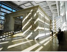 Interno di un museo moderno con ampie vetrate e soffitto a griglia, che ospita una grande struttura in pietra scolpita con bassorilievi e una scalinata d’accesso. La luce naturale crea lunghe ombre geometriche sul pavimento lucido.